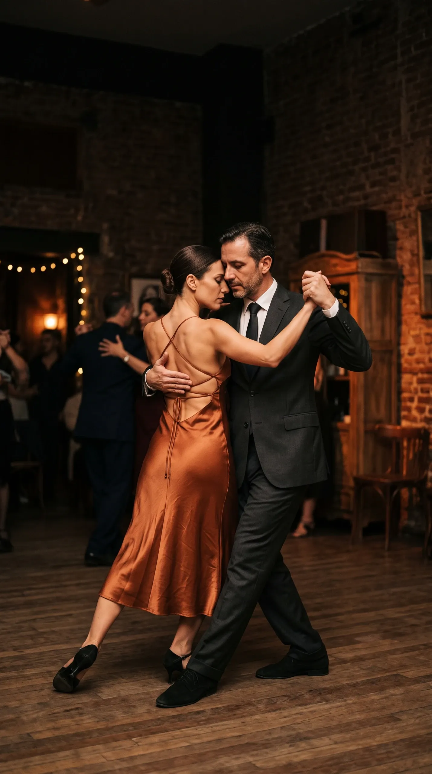 A man in a dark charcoal suit and a woman in a backless copper satin midi dress dance Argentine tango in an intimate embrace on a polished wooden floor in a rustic, atmospheric venue with exposed brick and warm background lighting.