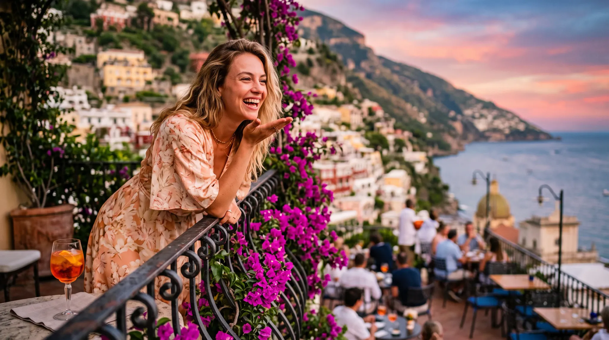 A vibrant photograph of a laughing woman leaning over a decorative wrought-iron balcony railing intertwined with blooming magenta bougainvillea. Dressed in a light floral wrap dress with delicate gold jewelry, she gestures happily with an iced orange spritz cocktail resting on the balustrade beside her. In the soft sunset background, pastel buildings of a coastal Mediterranean cliffside village cascade down to the sea, with diners seated on a restaurant terrace below.