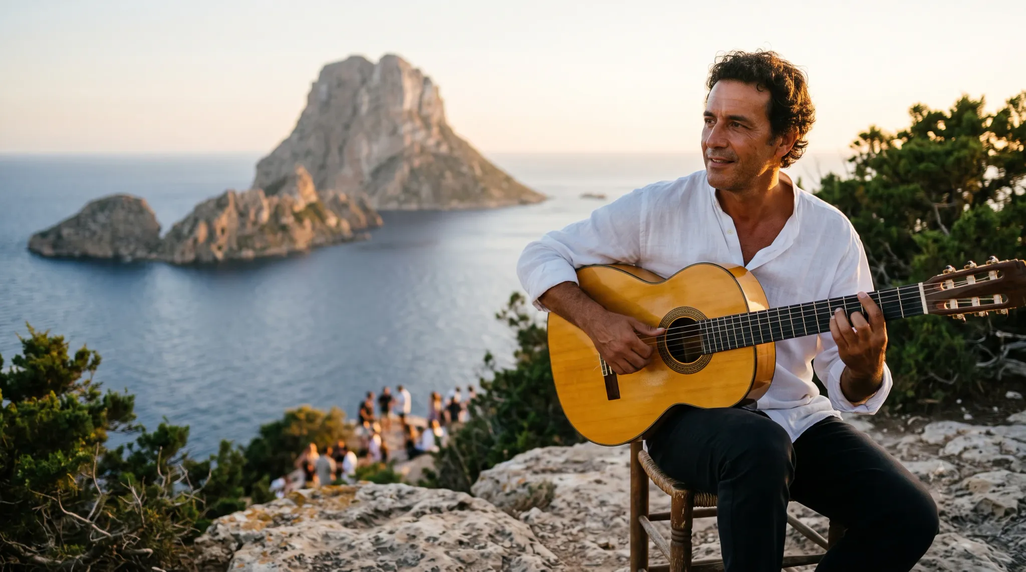 A middle-aged man with curly dark hair sits on a wooden stool on a rocky cliff edge, playing a classical acoustic guitar with evident focus and a subtle smile. He wears a loose white linen-looking long-sleeve shirt with the collar open and dark trousers. Behind him stretches a calm blue sea with a dramatic rocky sea-stack island rising in the distance, bathed in warm golden sunset light. Green shrubs and trees frame both sides. A small group of people is visible further below on the clifftop, gathered to watch or enjoy the view. The scene conveys warmth, artistic performance, and natural grandeur.