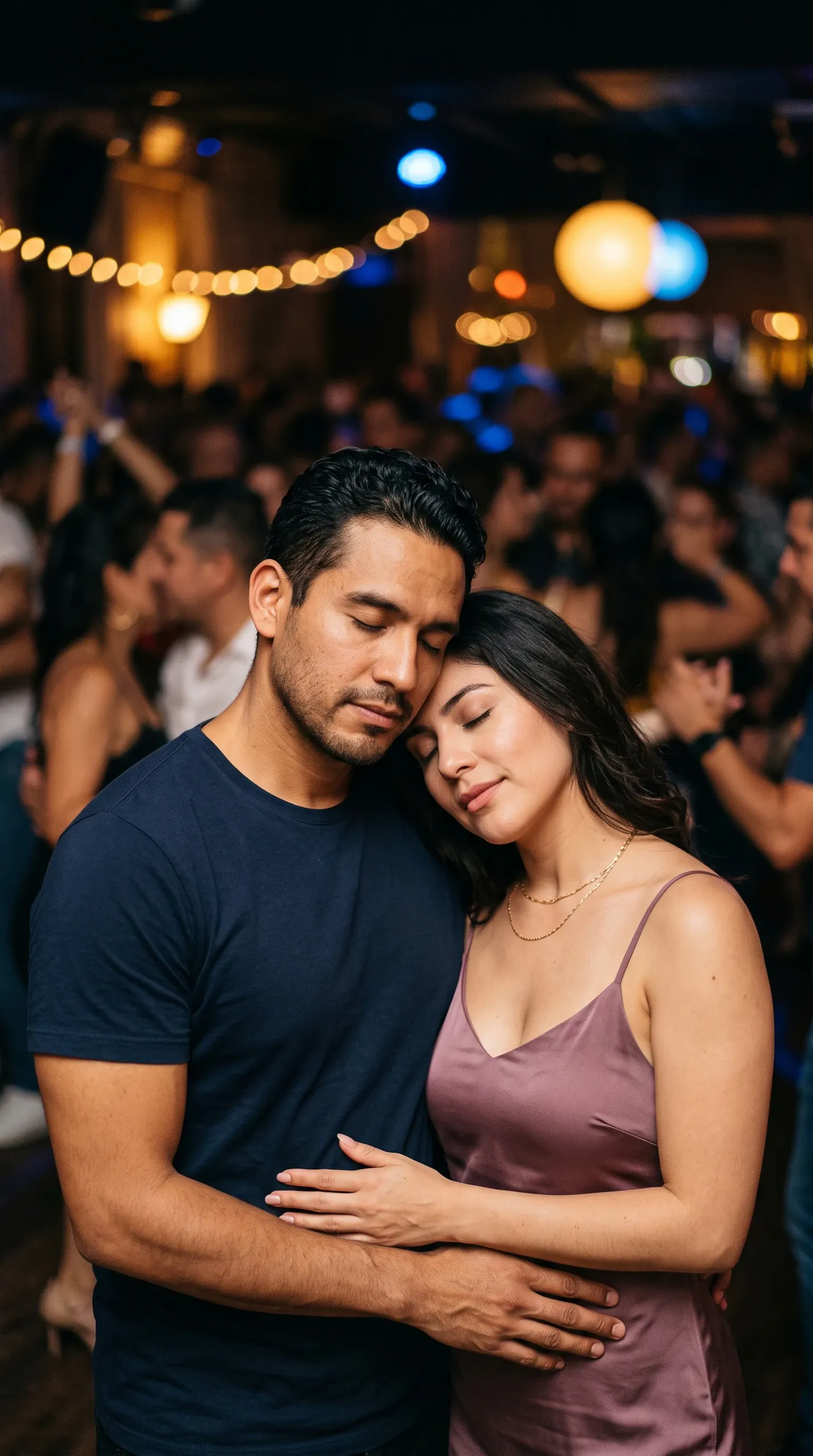 A close-up waist-up portrait of a man and woman dancing intimately with their eyes closed in a crowded indoor dance venue or nightclub under warm string lights. The man wears a navy blue t-shirt with his arms wrapped around the woman's waist, while the woman, dressed in a mauve slip dress and delicate gold chain necklace, rests her head on his chest and shoulder.