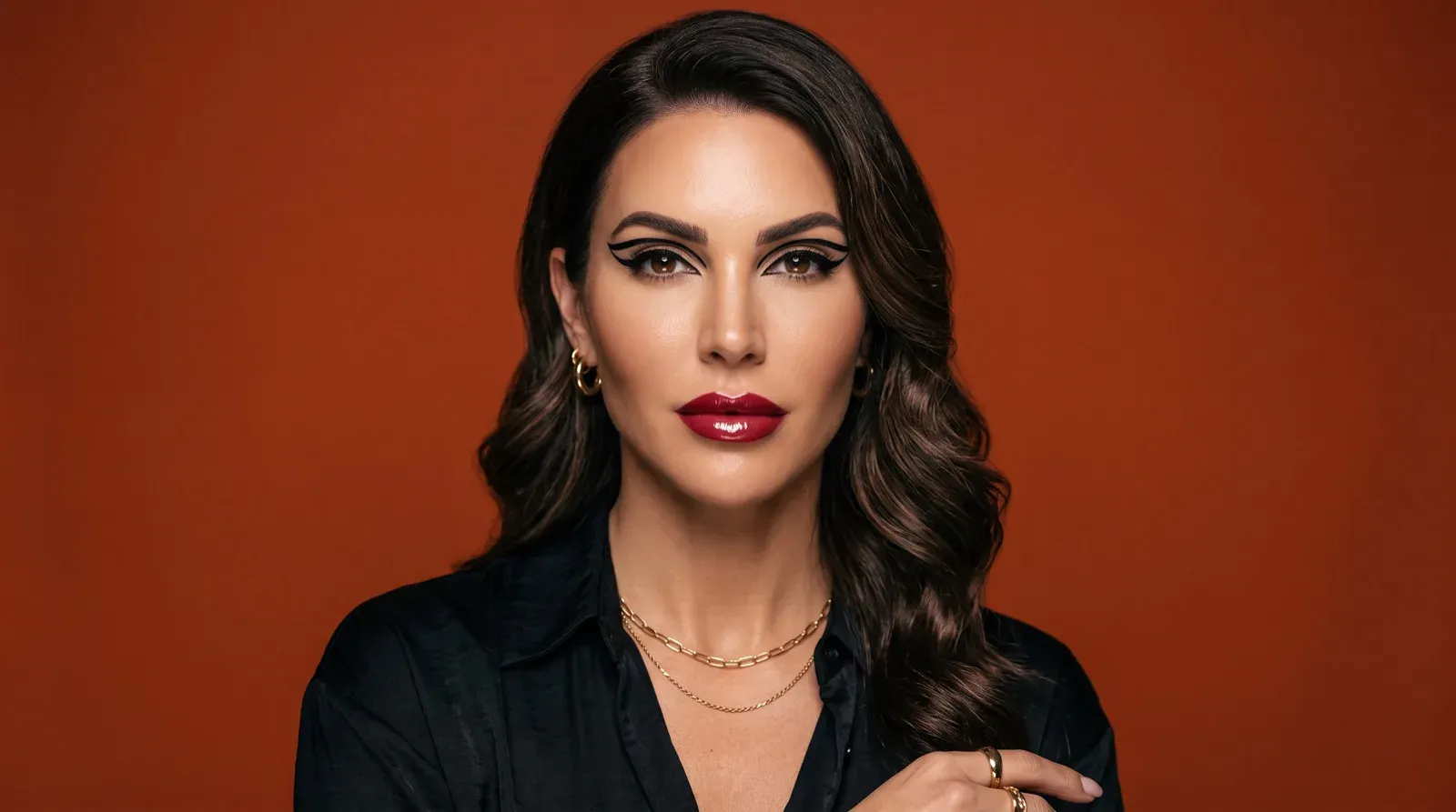 A studio portrait of an adult woman with graphic black winged eyeliner, glossy red lipstick, and dark wavy hair, wearing a black collared shirt, gold hoop earrings, and layered gold chain necklaces against a solid terracotta backdrop.