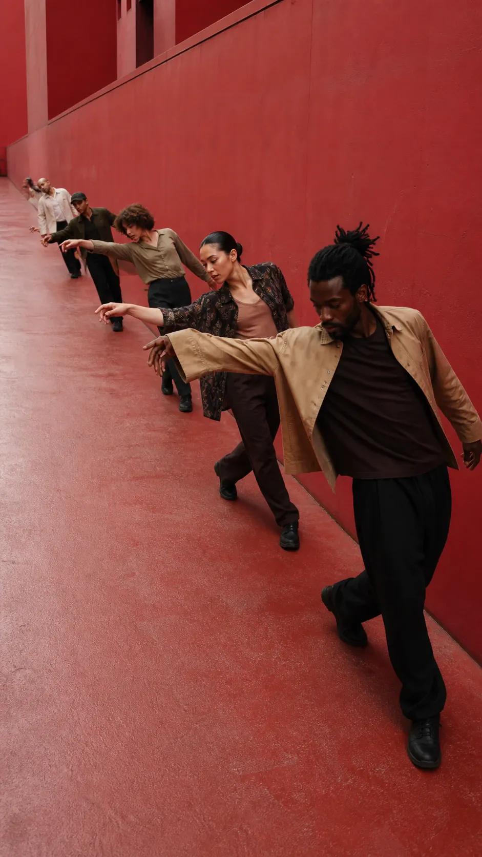 A line of six dancers performs a synchronized, leaning movement along a monochrome red corridor. The foreground dancer wears a tan overshirt over a dark shirt with loose black trousers and black shoes; behind him, dancers in patterned, brown, olive, and light clothing extend their arms in staggered formation. The image is a vertical, wide-angle color photograph with generous red floor and wall negative space.