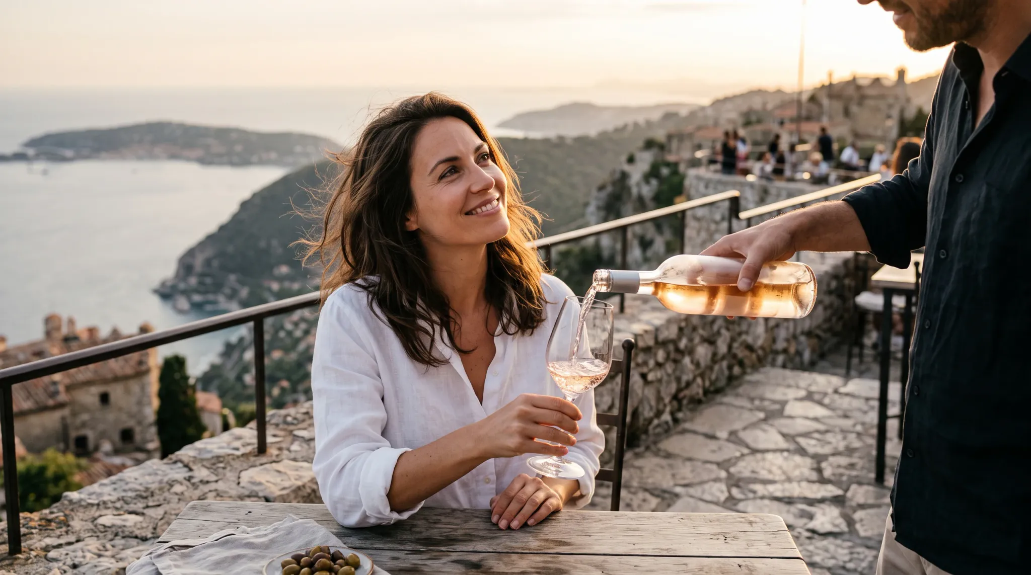 A smiling woman in a white linen shirt sits at an outdoor wooden table on a coastal terrace at sunset as a companion pours chilled rosé wine into her glass. In the background, rolling Mediterranean hills, traditional stone architecture, and the sea are softly illuminated by golden evening light.