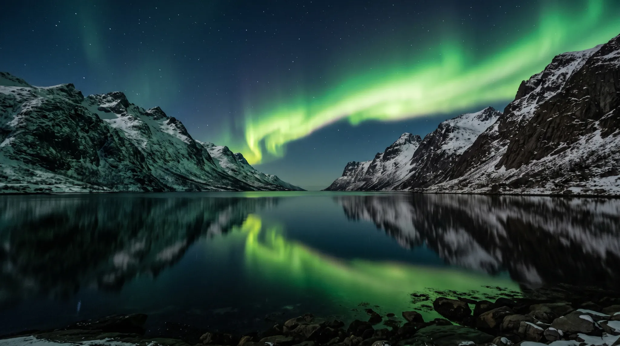A wide-angle nighttime photograph of the vibrant green Aurora Borealis arcing across a starry Arctic sky, set above dramatic snow-capped mountains and perfectly reflected in the calm waters of a coastal fjord with a rocky foreground shore.