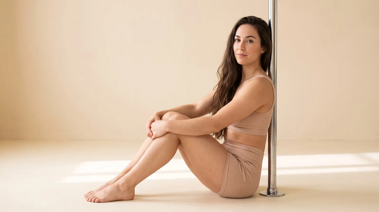 A woman with long wavy brown hair sits barefoot on the floor of a minimalist studio in a ribbed beige sports bra and matching shorts, leaning her head and back against a vertical chrome dance pole while looking forward.