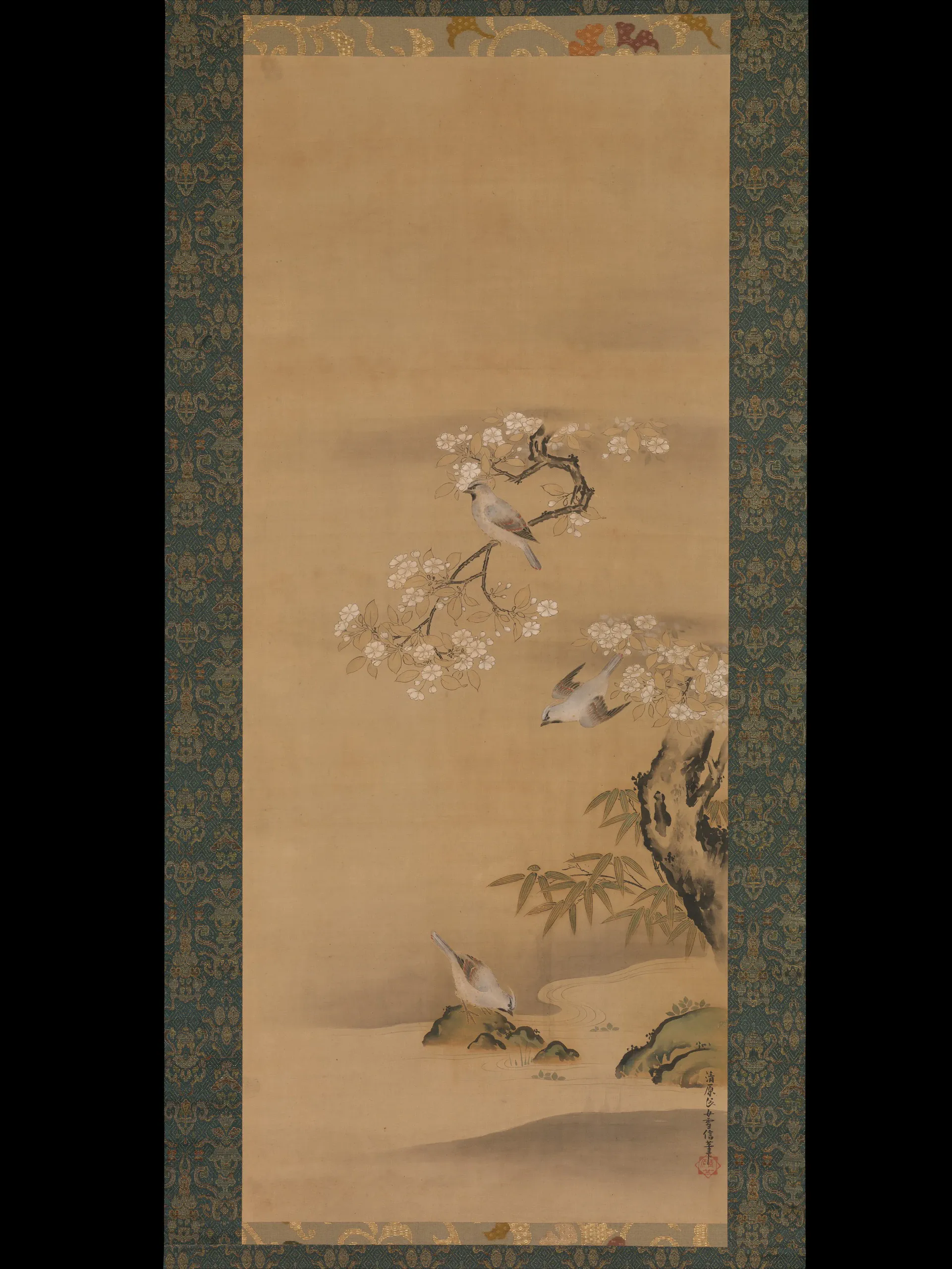 A vertically framed East Asian-style hanging-scroll painting shows three small birds among flowering branches, bamboo, rocks, and a calm water feature. One bird perches centrally on a blossom branch, another flies at right, and a third stands near the water below. The painting has a warm beige field, dark patterned blue-green textile borders, calligraphic marks, and a red seal.