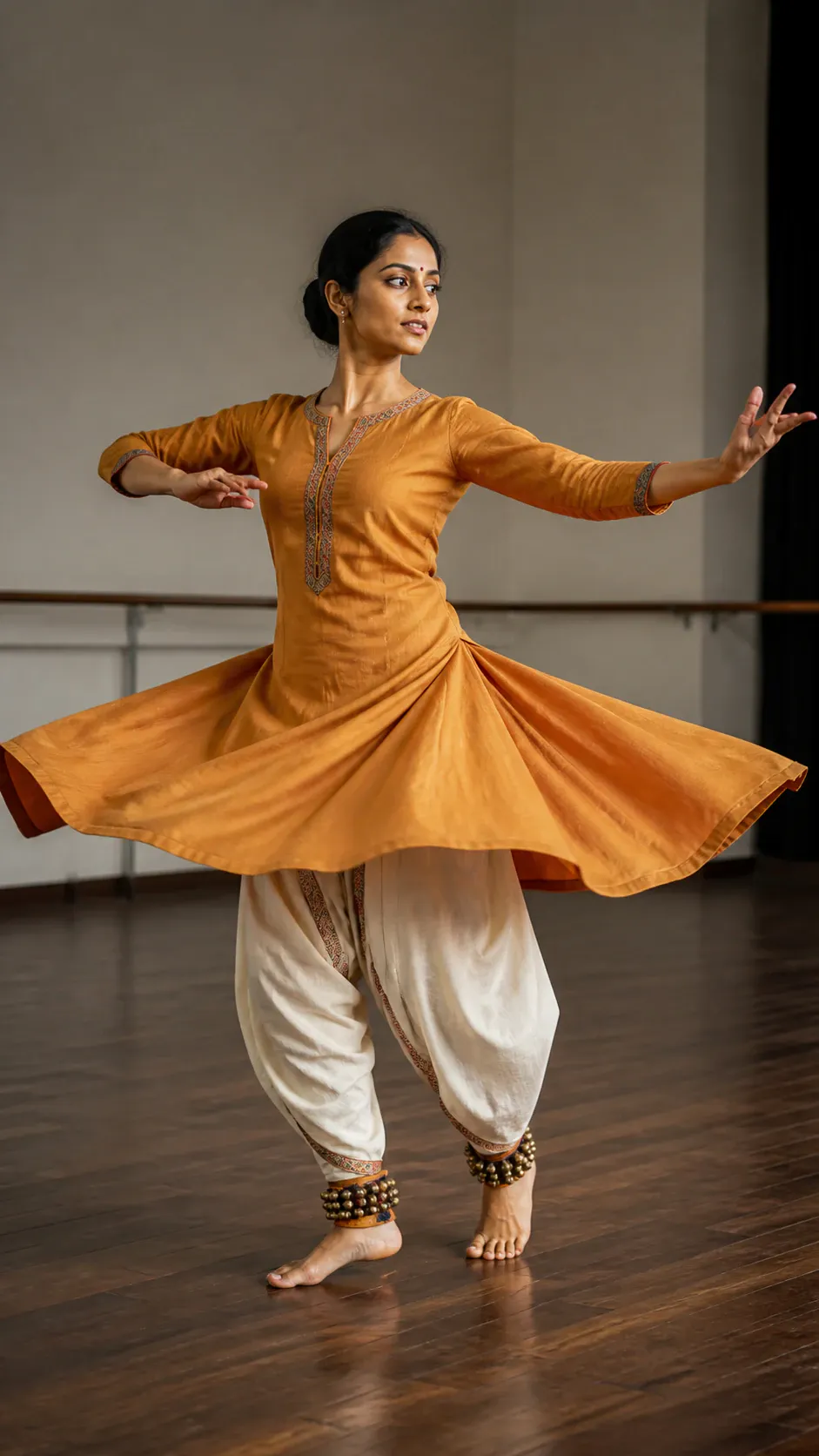 A single adult female dancer performs in an indoor dance studio, extending both arms while her orange long-sleeved tunic flares outward over loose white trousers. She wears ankle bells and dances barefoot on a polished wooden floor, with a barre visible along the rear wall.