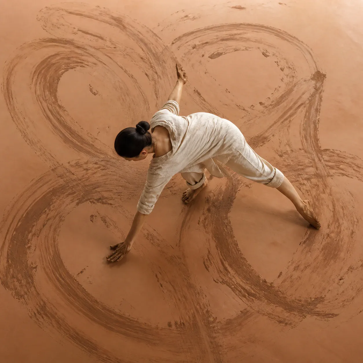 A single adult woman in a loose off-white long-sleeve top and matching wide-leg trousers holds a dynamic floor-based stretching pose, with one knee bent, one leg extended sideways, and both arms reaching diagonally across a warm terracotta studio floor marked by broad circular brown pigment or brush strokes. Her dark hair is tied in a high bun, and she is barefoot. The image is a square, top-down full-body composition with soft warm light and generous surrounding floor space.
