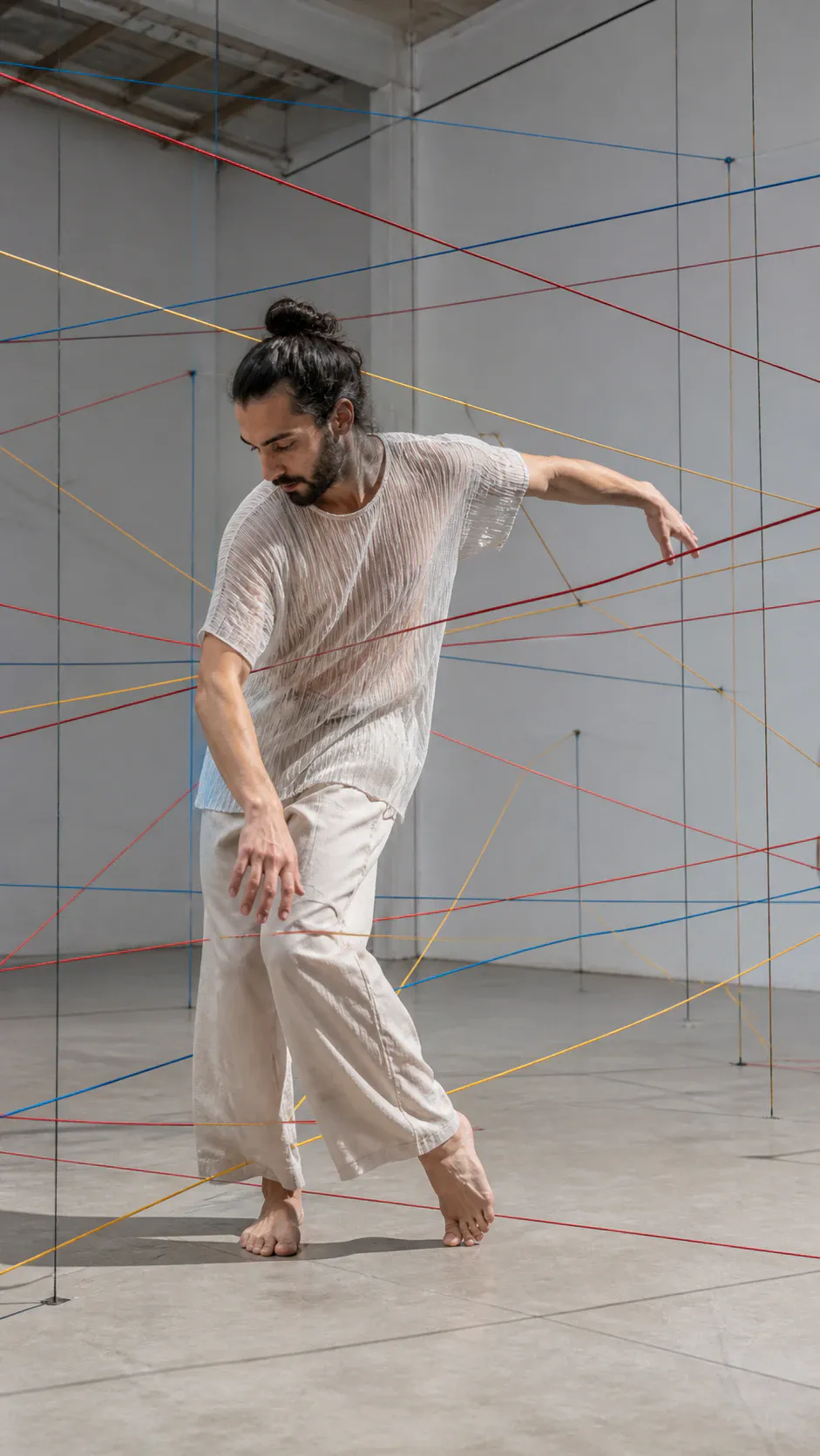 A barefoot male dancer with dark hair tied in a bun and a beard performs expressive, fluid movements amidst a complex geometric web of colorful taut strings crisscrossing an airy white studio or gallery room. He wears a sheer, crinkled short-sleeved white tunic top and relaxed wide-leg beige linen pants.