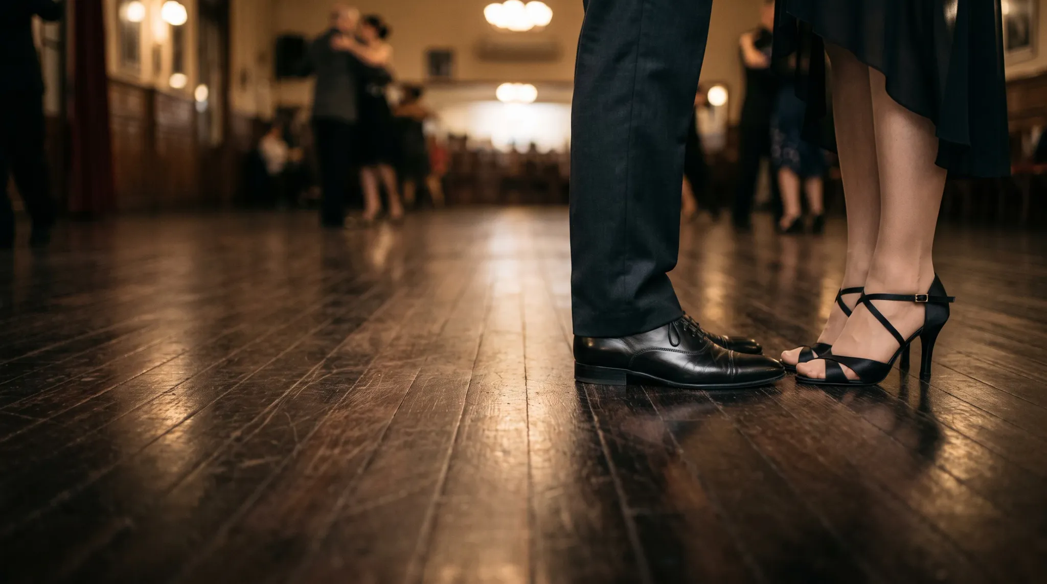 A low-angle, shallow depth of field photograph focusing on the feet and lower legs of a couple dancing in a ballroom. The man wears dark trousers and polished black leather dress shoes, while the woman wears a dark dress and black open-toe strappy high-heeled dance sandals. Other dancing couples are visible in the warm, softly blurred background on the polished wooden floor.