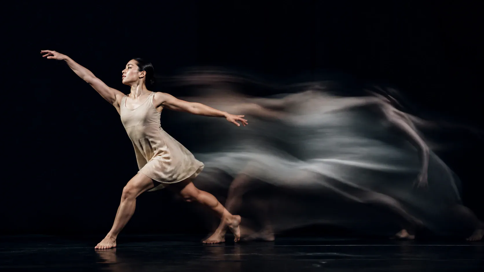 A female contemporary dancer in a beige sleeveless dress performs an expressive movement across a dark stage, captured with a dynamic long-exposure motion blur trail trailing behind her sharp primary figure.
