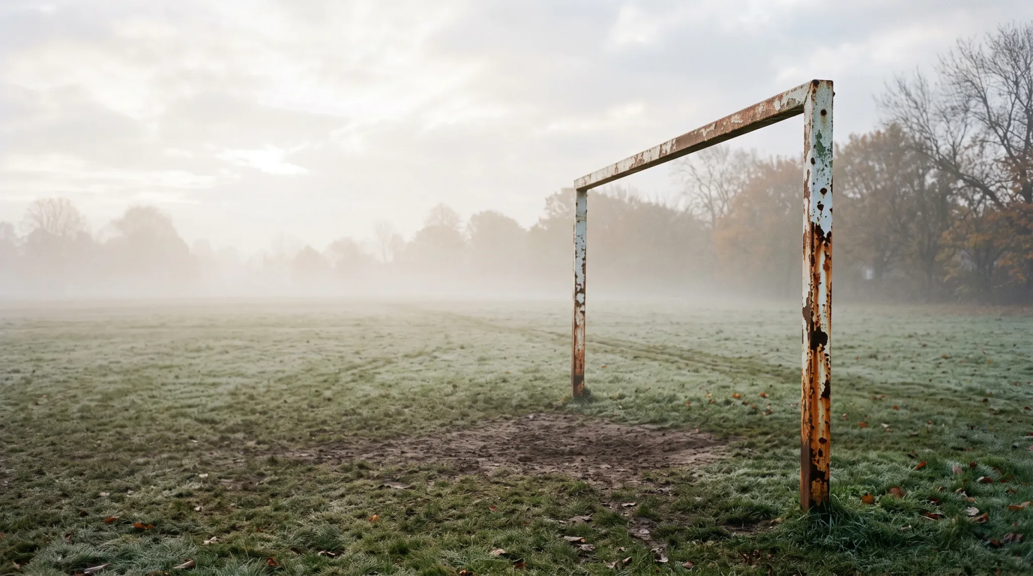 A rusty, peeling white metal goal post stands in a wide, misty grass field covered with light morning frost. A worn patch of bare mud lies directly under the goalmouth, while distant trees fade into dense fog under an overcast sky.