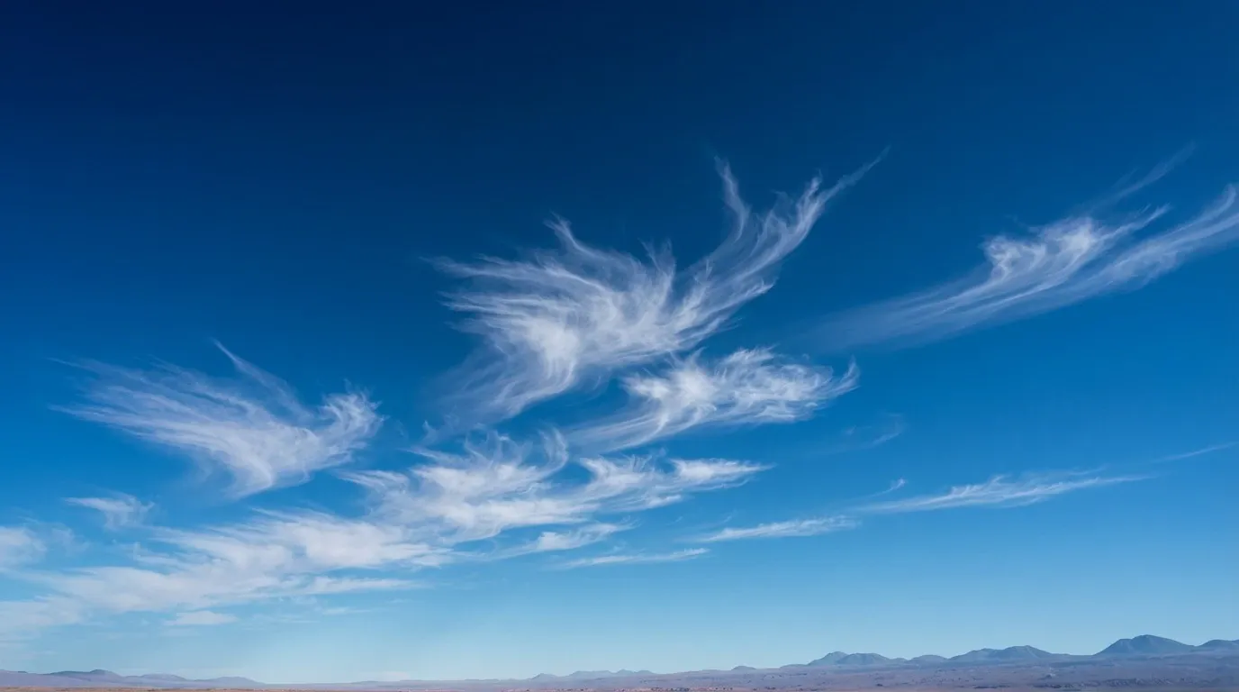 A wide landscape photograph capturing delicate, feather-like cirrus cloud formations swirling across a vibrant deep blue sky. Along the low horizon in the far distance, an arid desert plain stretches beneath distant mountain silhouettes under bright natural sunlight.