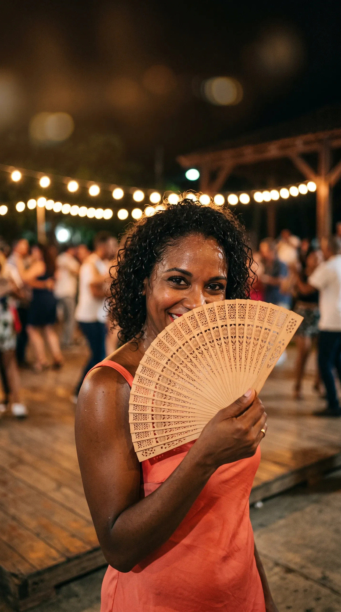 A vibrant nighttime portrait of a smiling woman holding an intricate light-colored wooden folding hand fan partially covering the lower half of her face. She has curly dark hair, radiant warm skin, and wears a sleeveless coral top or dress along with a gold band on her ring finger. Warm overhead string lights illuminate the outdoor venue where a lively crowd dances in the softly blurred background on a wooden platform.
