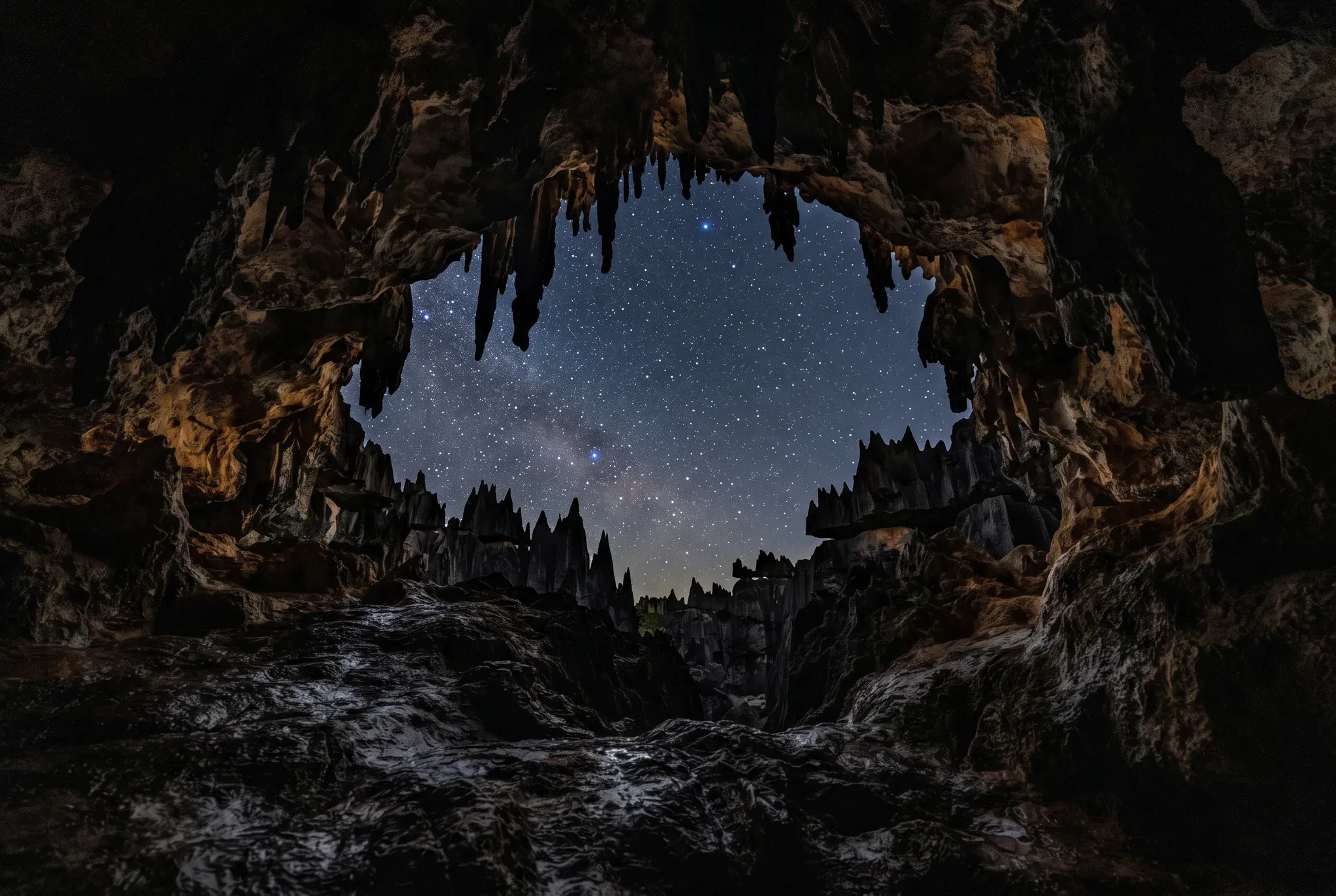 A wide-angle night photograph taken from inside a textured limestone cave looking out through its natural archway opening. Sharp stalactites hang from the cave ceiling, framing a dark sky filled with countless stars and celestial dusting, with jagged karst limestone pinnacles rising in the middle distance.
