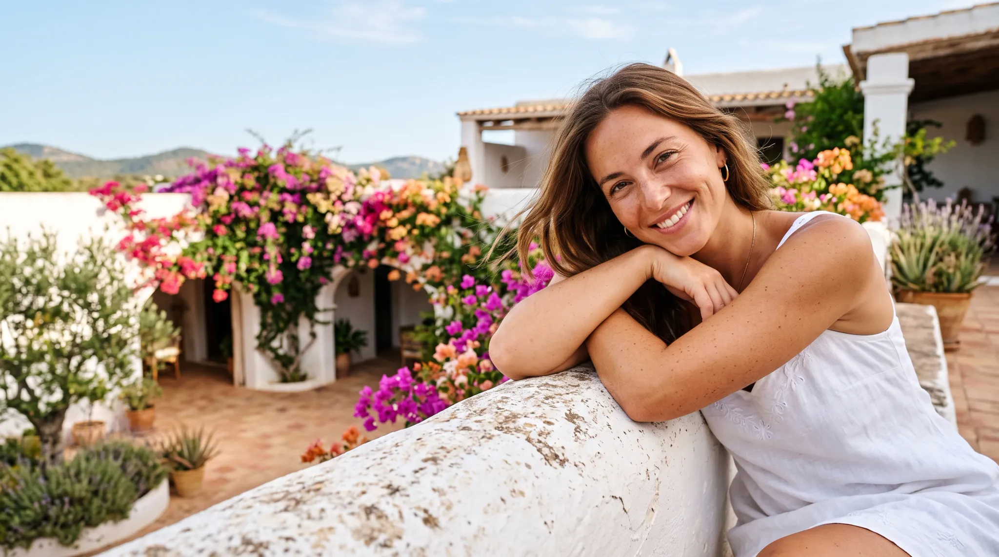 A close waist-up shot of a joyful young woman leaning her crossed arms on a weathered whitewashed stone balustrade on an elevated Mediterranean terrace. She wears a white sleeveless dress, gold hoop earrings, and a delicate gold necklace while smiling broadly at the camera. In the soft-focus courtyard below, vibrant magenta and orange bougainvillea frame traditional white stucco architecture under a bright, sunny sky.