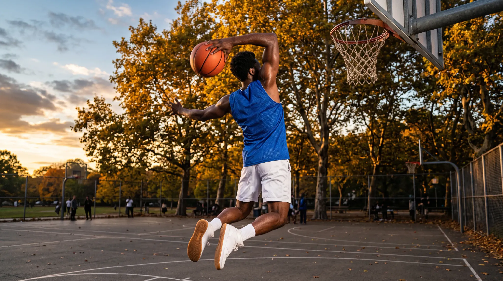 A muscular male basketball player in a royal blue mesh tank top and white shorts is captured in mid-air during a dynamic leap towards a basketball hoop on an outdoor court. He holds an orange basketball overhead with his right hand against a background of autumn trees illuminated by golden hour sunlight.