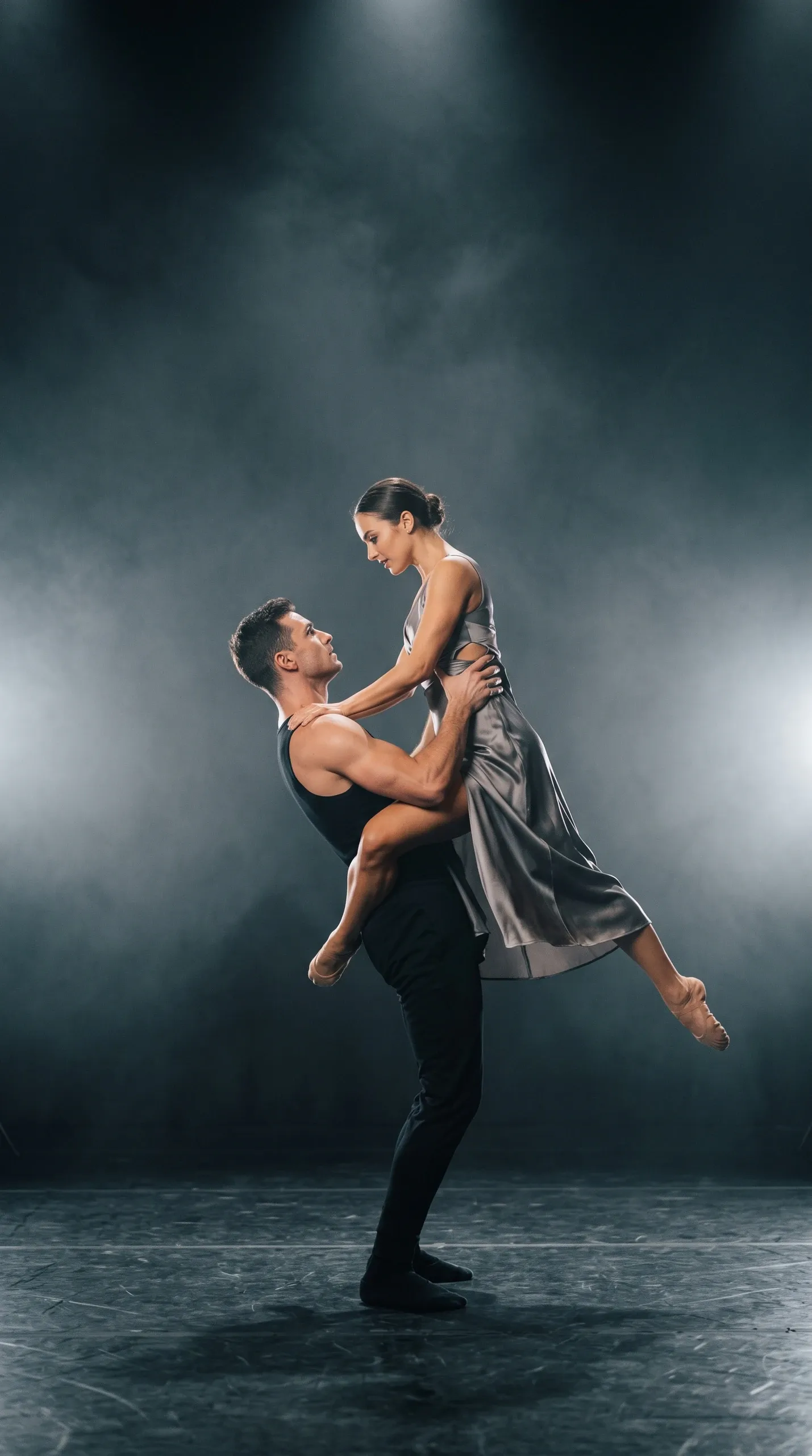 Full-length view of two contemporary dancers in a dramatic partnered lift on a dark, hazy stage. The male dancer, dressed in a black sleeveless tank top and fitted trousers, stands upright and holds the female dancer suspended in mid-air. She wears a sleeveless silver-gray satin midi dress with side cutouts and soft beige dance flats, arching her body with legs positioned gracefully as they gaze intently at each other under strong theatrical rim lighting.