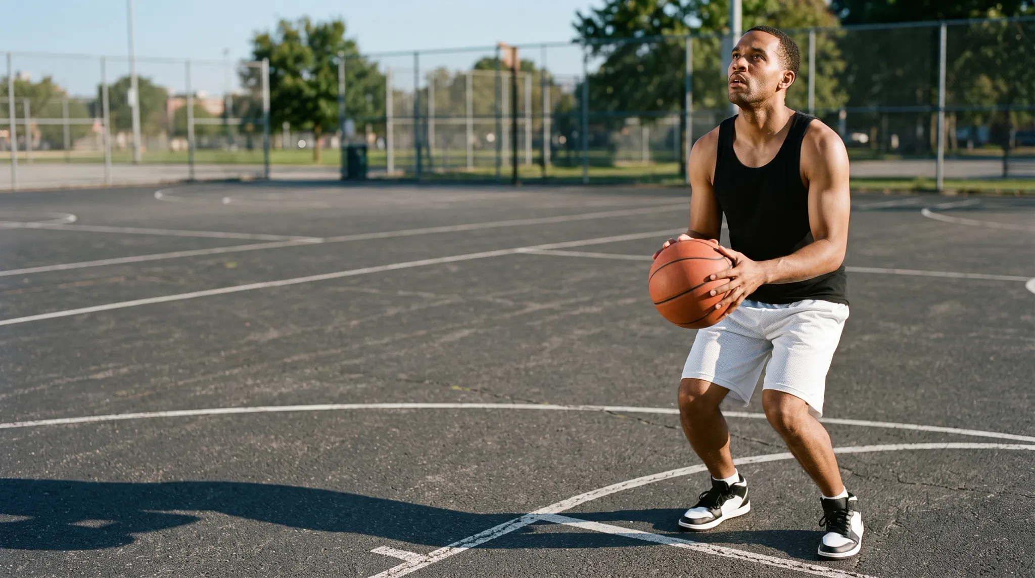 A man in a black tank top, white shorts, and black-and-white sneakers holds an orange basketball in a partial squat on an outdoor asphalt basketball court. He is positioned on the right side of the wide frame and looks upward, with fenced courts, grass, and trees in the background.