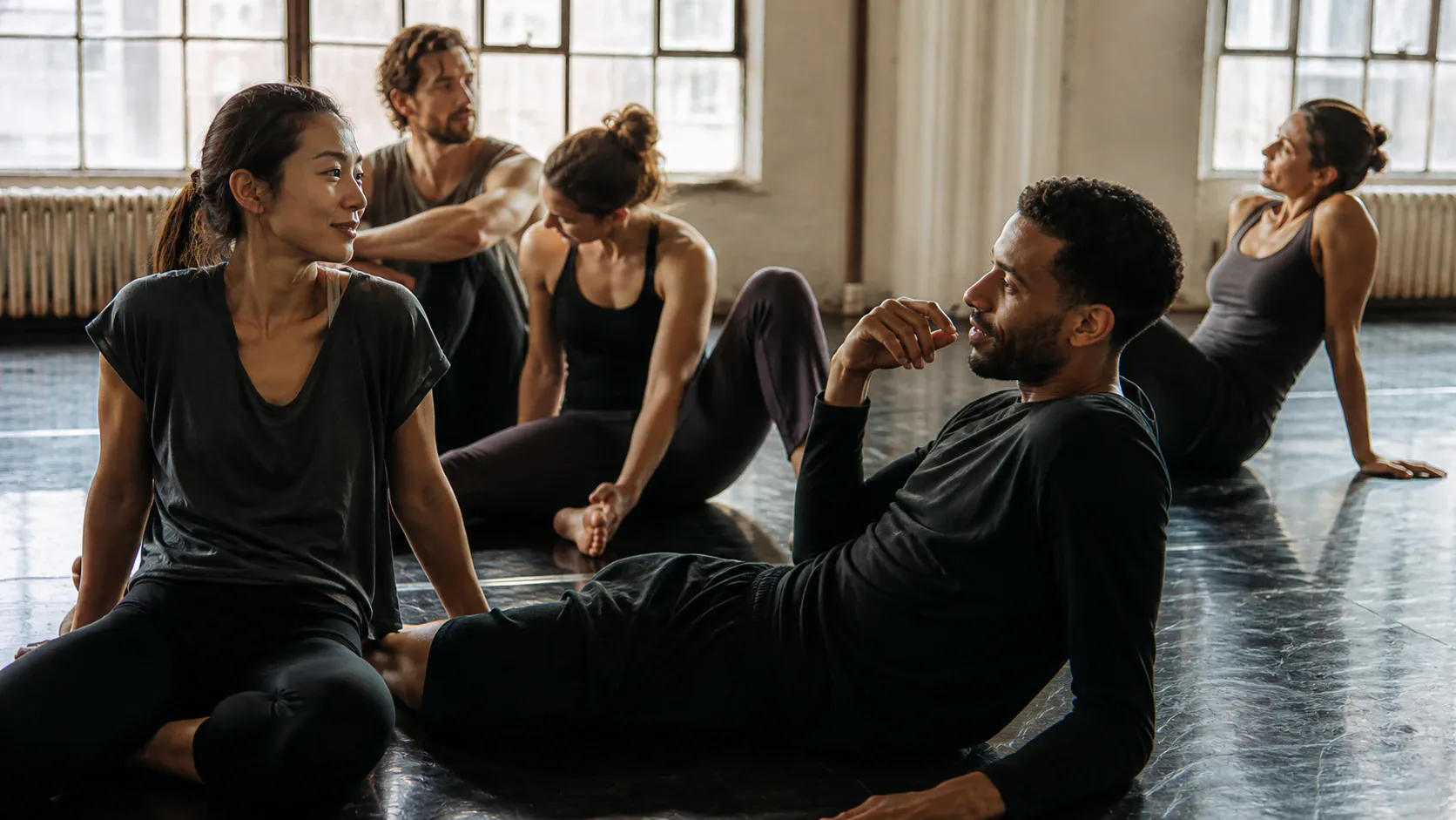 A group of five adult dancers or athletes rest on the reflective dark floor of a bright, sunlit rehearsal studio with expansive gridded windows. In the foreground, a seated woman and a reclining man in black athletic wear engage in conversation. Three other individuals relax in various seated poses across the room.