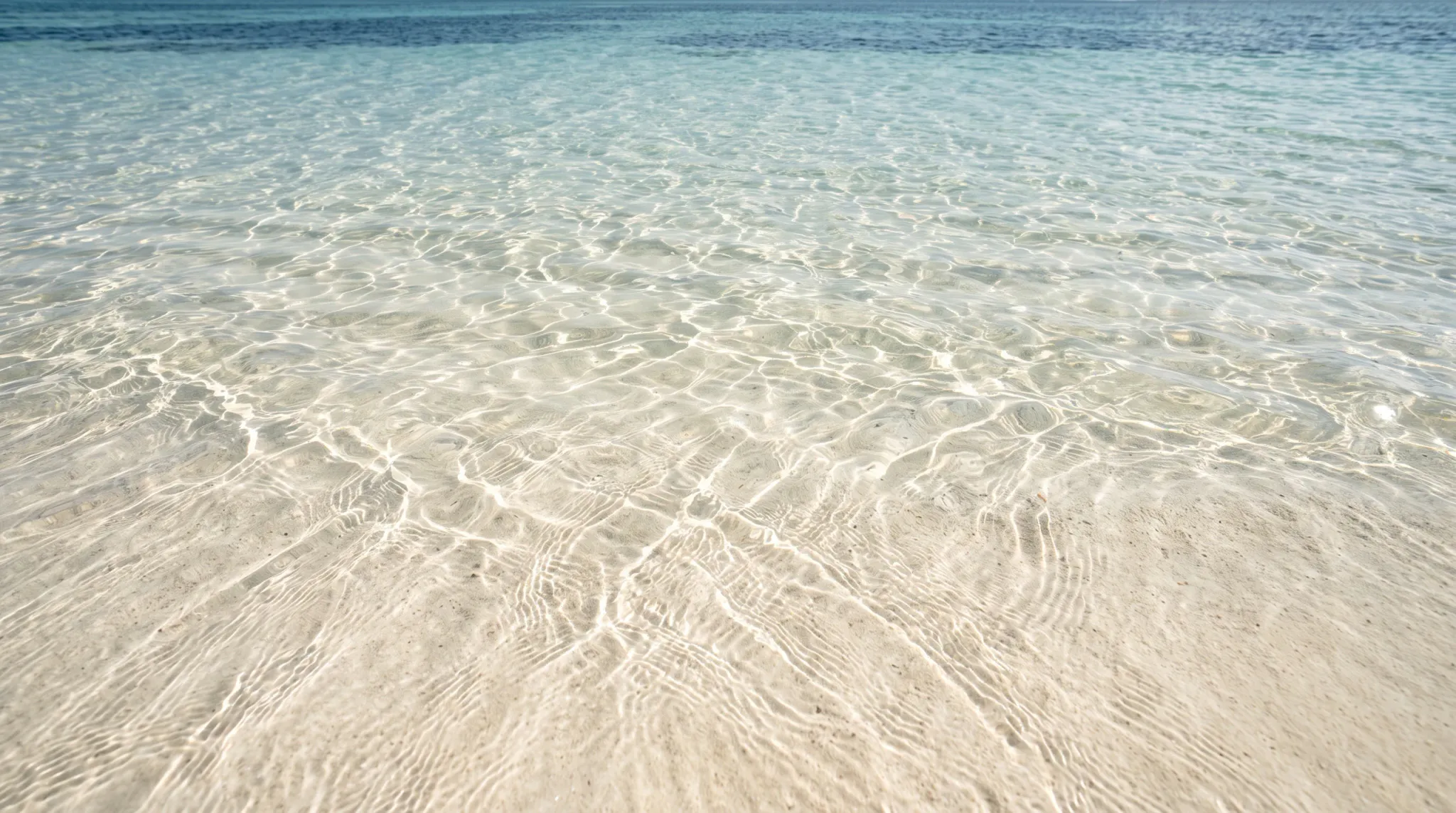A high-angle photograph capturing crystal-clear, shallow turquoise and aqua ocean water gently rippling over a pale, fine sand seabed. Direct bright overhead sunlight creates intricate dancing caustic patterns and bright reflections across the submerged sand, while the upper background transitions into deeper blue coastal waters under a clear open environment.