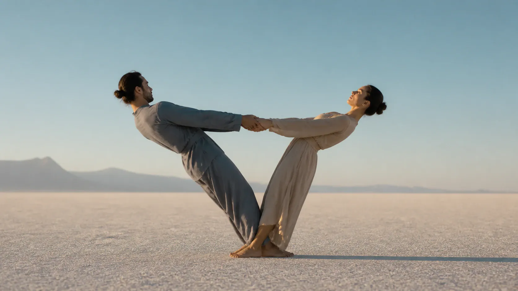 A man in gray and a woman in a flowing beige dress lean away from each other while holding hands in a synchronized balancing pose on a vast white salt flat. Distant mountain silhouettes line the horizon beneath a clear blue sky, with warm directional sunlight and generous open space.