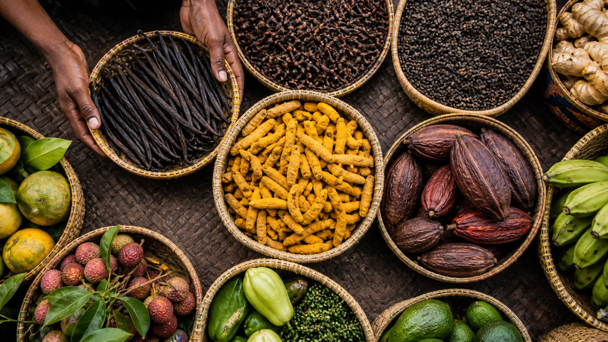 Baskets of spices, vanilla and tropical produce form a richly colored market still life.