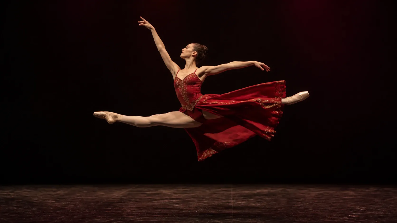 An adult ballerina in a sculptural crimson costume reaches the apex of a grand jeté above a matte black theatre floor, the skirt opening like a painted wing.